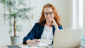 Photo of successful ginger female freelancer has remote work, watches webinar online on laptop computer, writes down information in notepad, smiles happily poses at workplace dressed in formal clothes